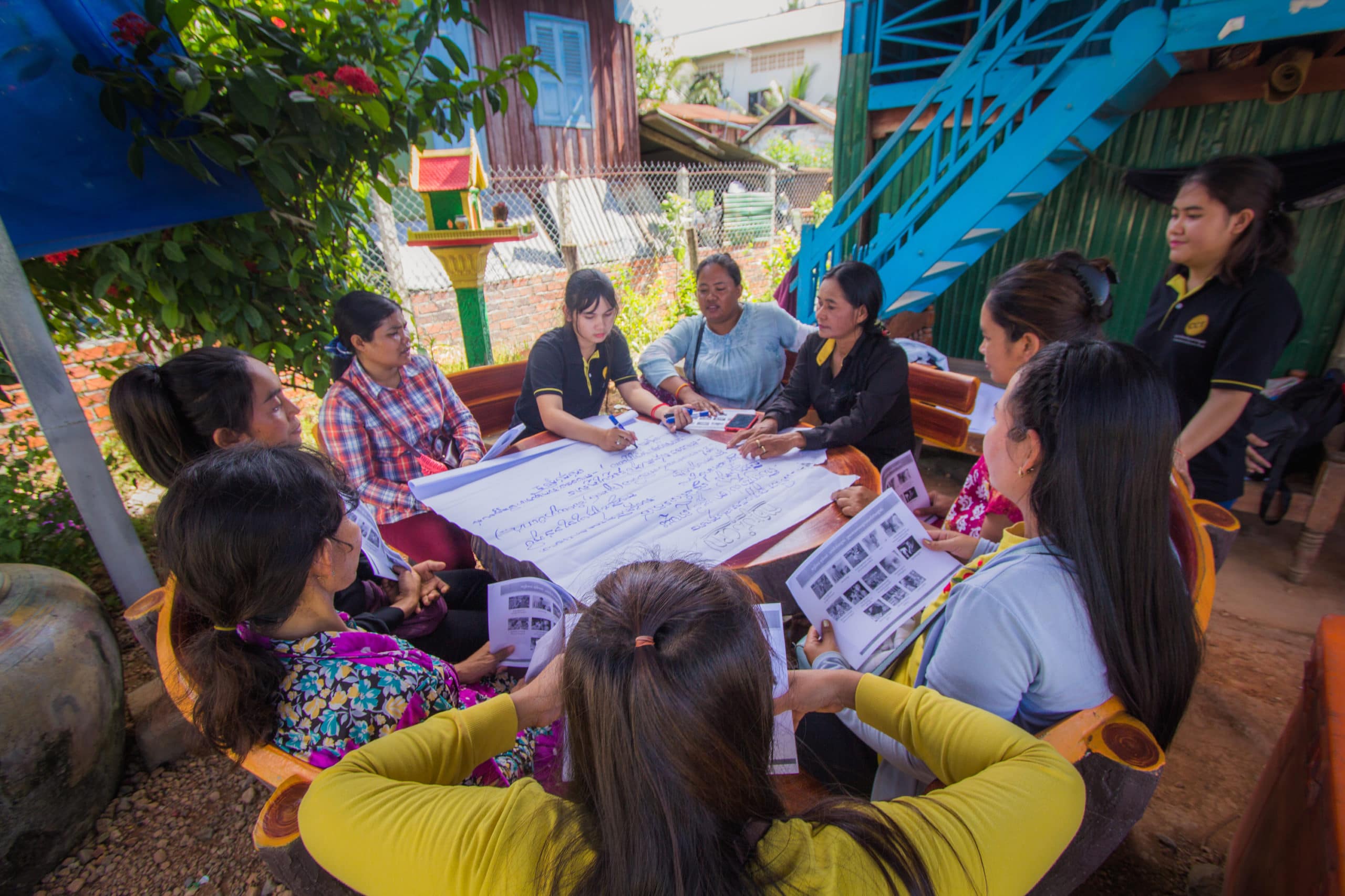CCT team members and women of a local Cambodian community gather around a round table  for a meeting. A large piece of paper covers the table and is covered in hand written notes